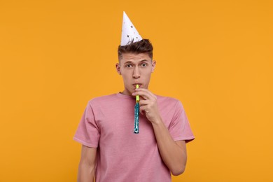 Young man in party hat with blower on orange background