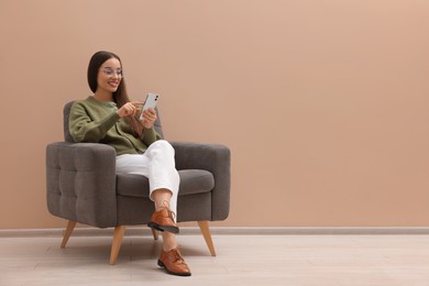 Photo of Beautiful woman using smartphone while sitting in armchair near beige wall indoors, space for text