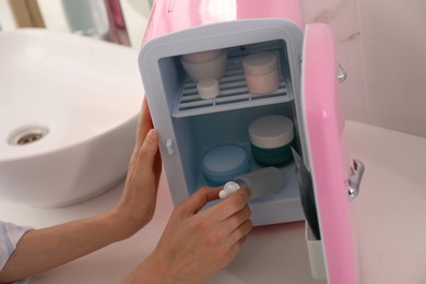 Photo of Woman taking cosmetic product from mini fridge at white table, closeup