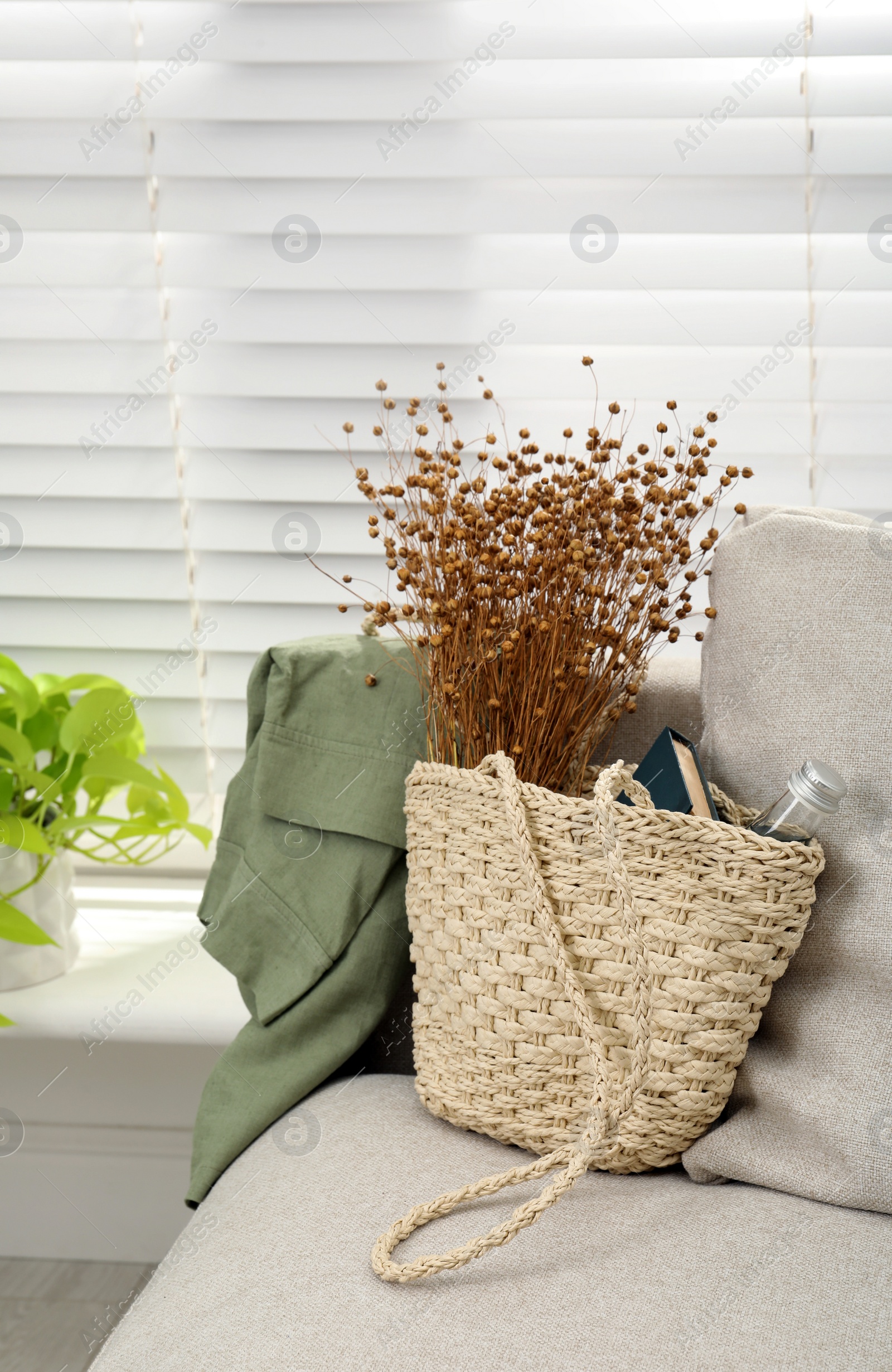 Photo of Stylish beach bag with wildflowers, bottle of water and book on sofa in room