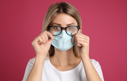 Woman wiping foggy glasses caused by wearing disposable mask on pink background. Protective measure during coronavirus pandemic