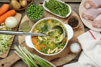 Saucepan of delicious vegetable soup with chicken and different ingredients on wooden table, flat lay