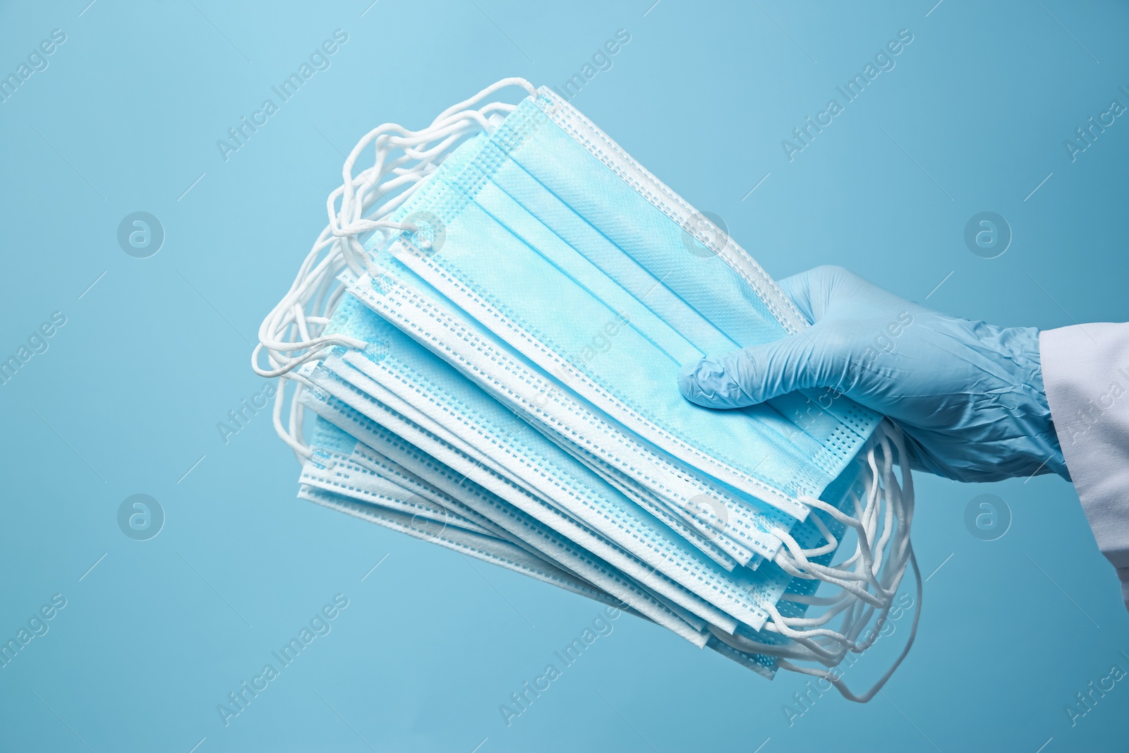 Photo of Doctor in latex gloves holding disposable face masks on light blue background, closeup. Protective measures during coronavirus quarantine