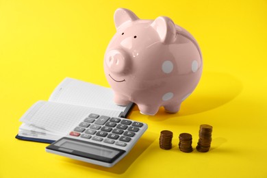 Calculator, pink piggy bank, notebook and stacked coins on yellow background