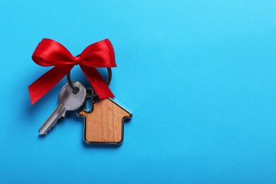 Photo of Key with trinket in shape of house and bow on light blue background, top view. Space for text. Housewarming party
