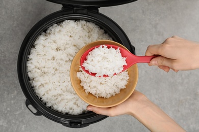 Photo of Woman putting boiled rice into bowl from cooker on grey background, top view