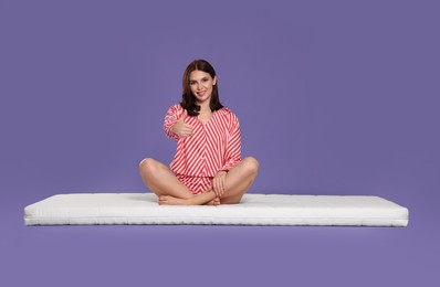 Young woman sitting on soft mattress and showing thumbs up against light purple background