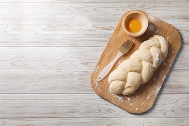 Photo of Raw braided bread, egg yolk and pastry brush on white wooden table, top view with space for text. Traditional Shabbat challah