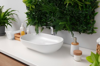 Photo of Green artificial plants, vessel sink and different personal care products in bathroom