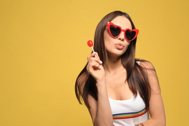 Portrait of beautiful young woman with heart shaped sunglasses and lollipop on color background. Space for text