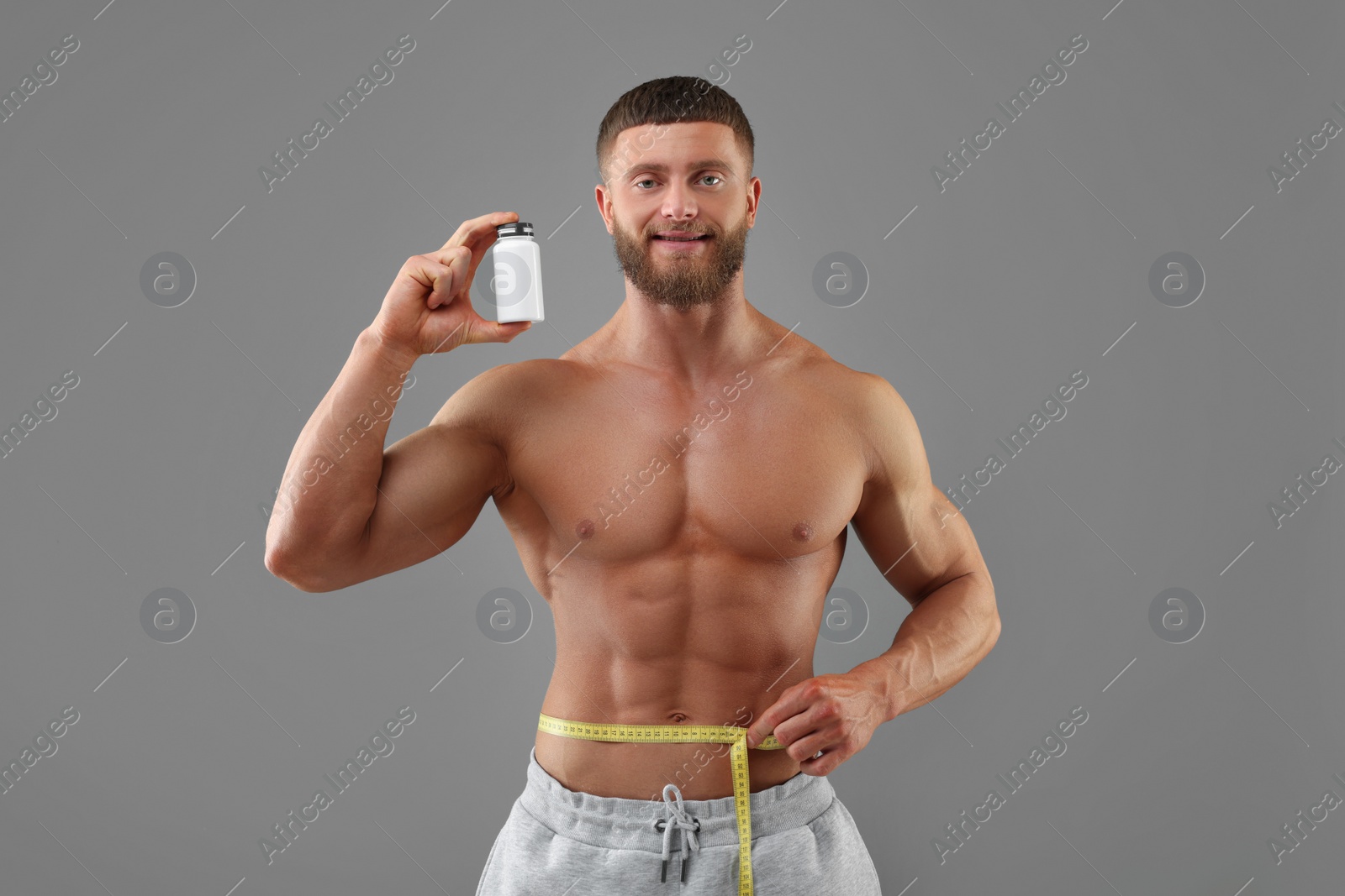 Photo of Athletic young man holding bottle of supplements and measuring his waist with tape on grey background. Weight loss