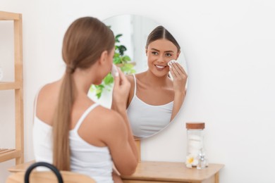 Photo of Beautiful woman removing makeup with cotton pad near mirror at home