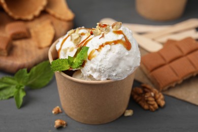Photo of Tasty ice cream with caramel sauce, mint and nuts in paper cup on grey wooden table, closeup