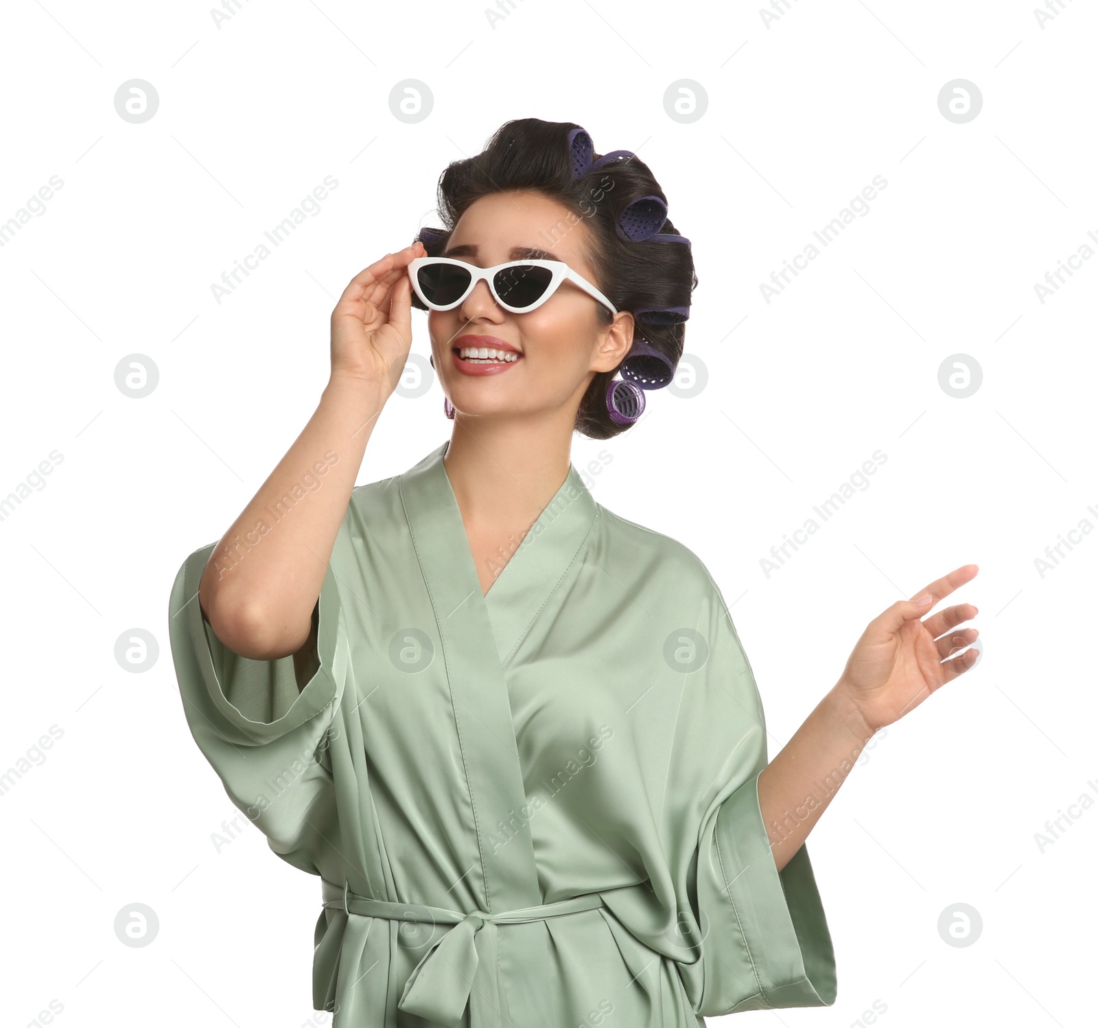 Photo of Happy young woman in silk bathrobe with hair curlers and sunglasses on white background