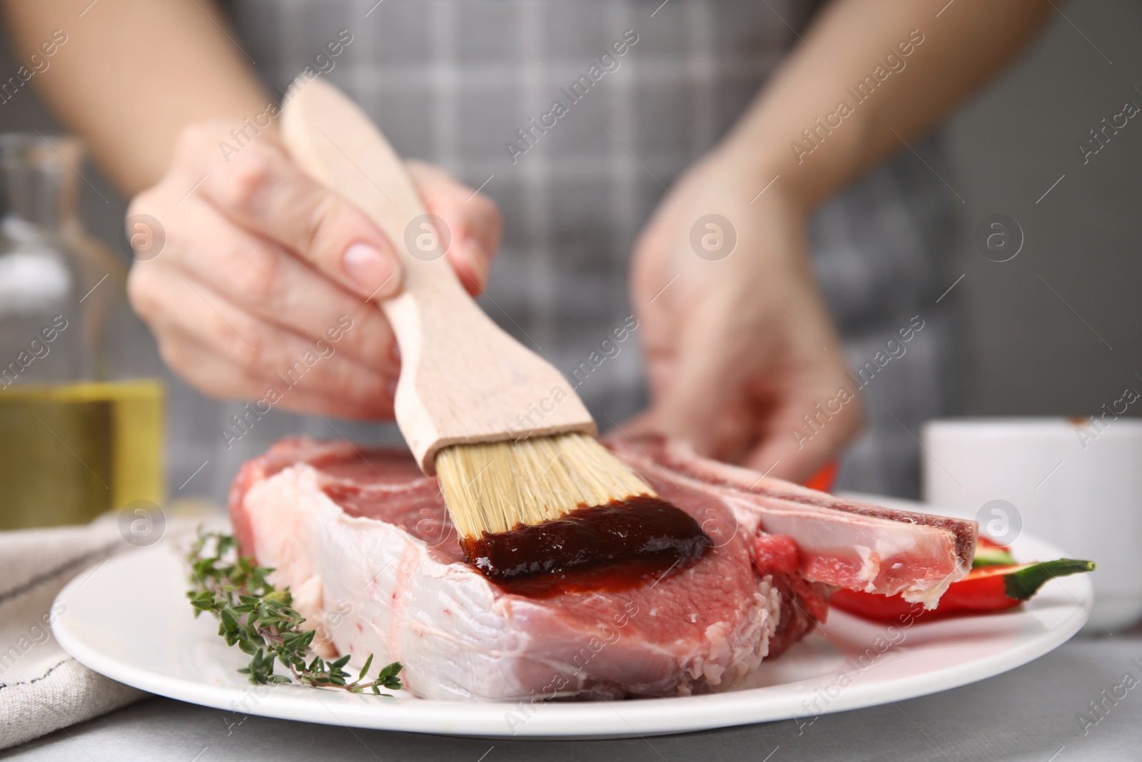 Photo of Woman spreading marinade onto raw meat with basting brush at table, closeup