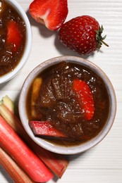 Photo of Tasty rhubarb jam in bowls, cut stems and strawberries on white wooden table, flat lay