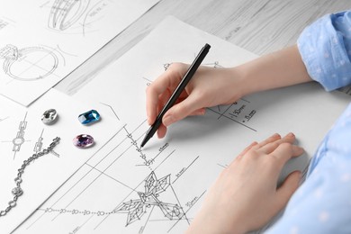 Photo of Jeweler drawing sketch of elegant necklace on paper at white wooden table, closeup