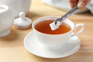 Woman with tongs adding sugar cube into cup of tea at wooden table, closeup