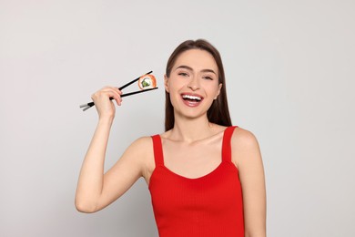 Photo of Happy beautiful young woman holding sushi roll with chopsticks on light background