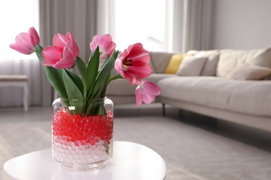 Photo of Different color fillers and tulips in glass vase on white table at home, space for text. Water beads