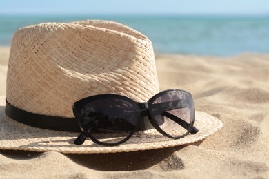 Photo of Hat with beautiful sunglasses on sand near sea, closeup