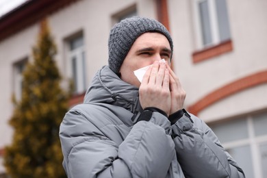 Photo of Sick young man with tissue blowing runny nose outdoors. Cold symptom