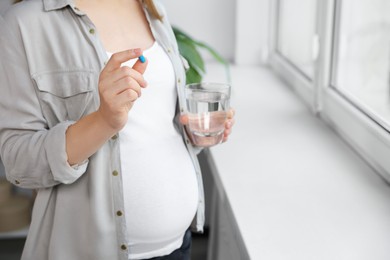 Photo of Pregnant woman holding pill and glass with water near window indoors, closeup