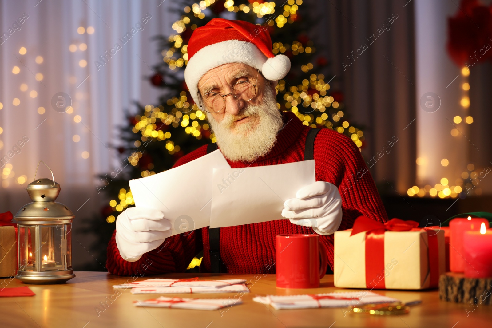 Photo of Santa Claus reading letters at his workplace in room decorated for Christmas