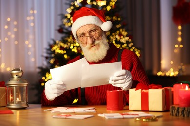 Photo of Santa Claus reading letters at his workplace in room decorated for Christmas