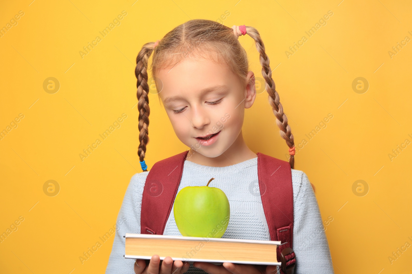 Photo of Cute little girl with apple, backpack and book on yellow background