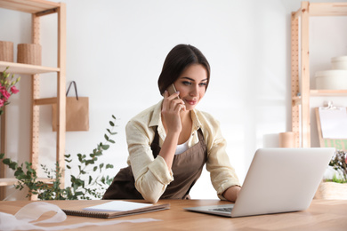 Photo of Florist talking on smartphone near laptop in workshop