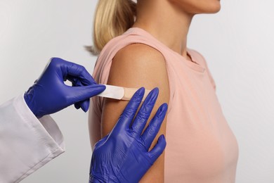 Nurse sticking adhesive bandage on woman's arm after vaccination on light background, closeup