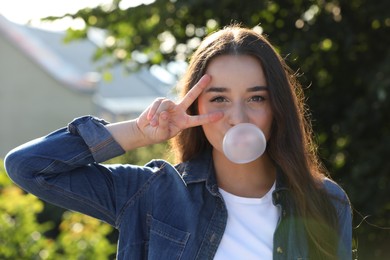 Beautiful young woman blowing bubble gum and showing V sign in park