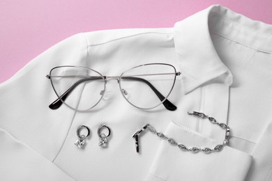 Stylish pair of glasses, jewelry and white blouse on pink background, flat lay