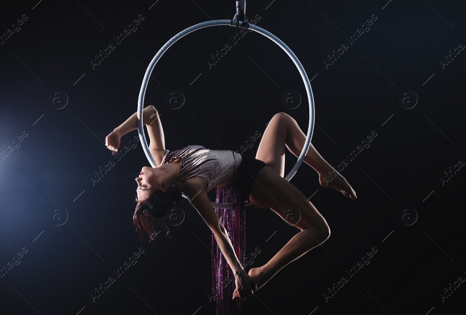 Photo of Young woman performing acrobatic element on aerial ring against dark background