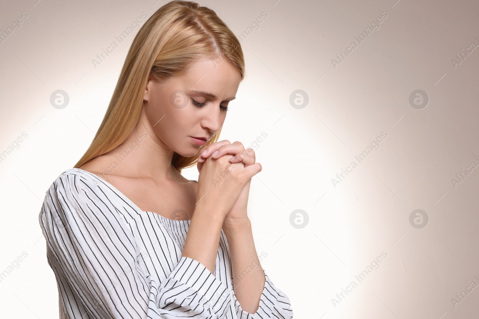 Photo of Religious young woman with clasped hands praying against light background. Space for text
