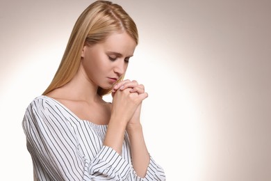 Photo of Religious young woman with clasped hands praying against light background. Space for text