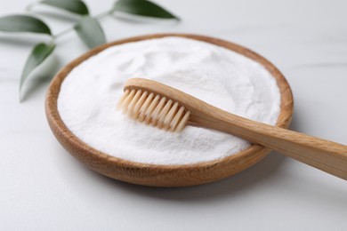Bamboo toothbrush, green leaf and bowl of baking soda on white marble table, closeup