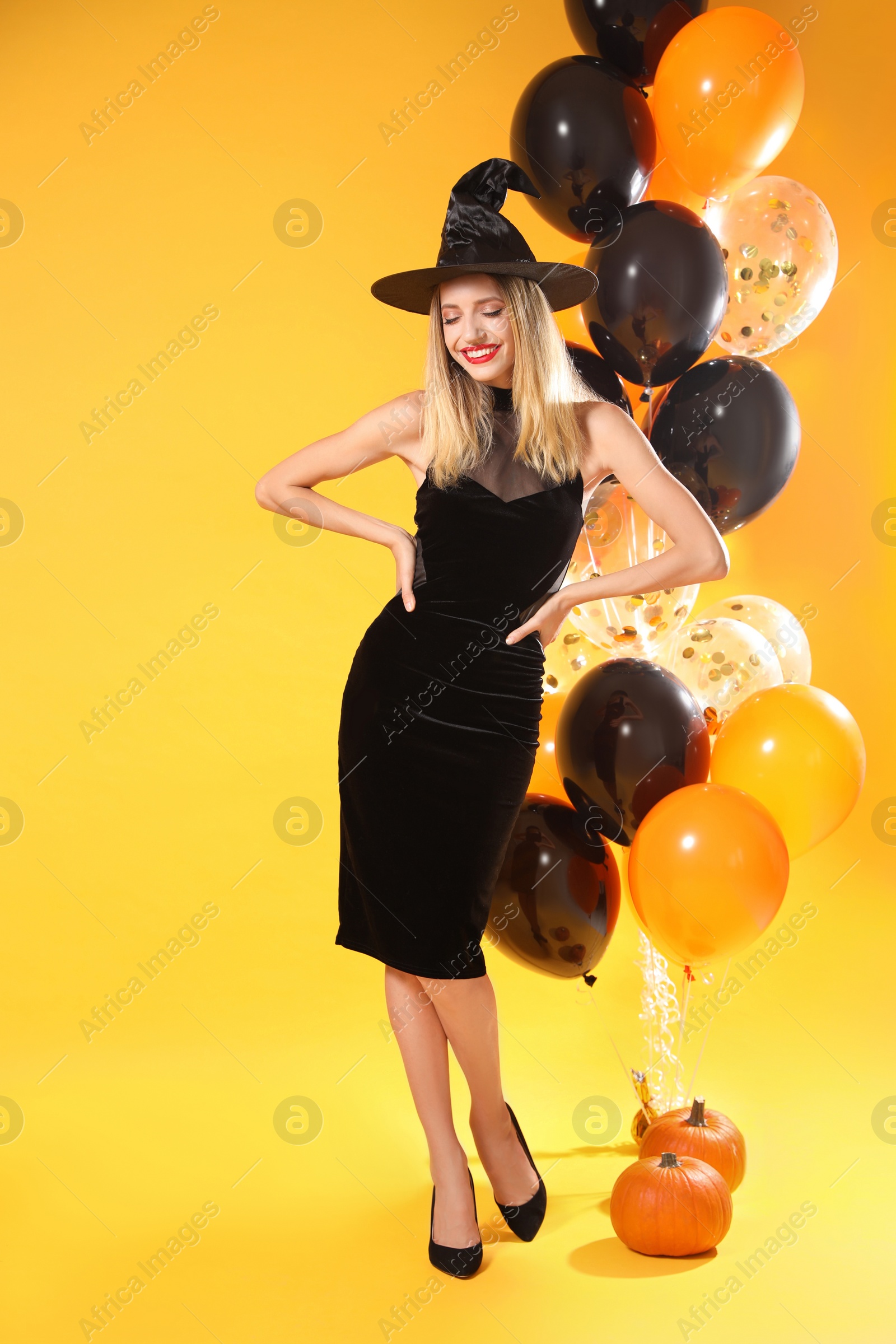 Photo of Beautiful woman in witch costume with balloons and pumpkins on yellow background. Halloween party