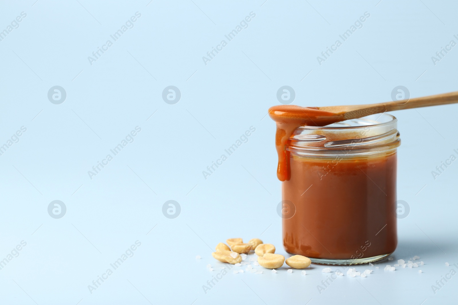 Photo of Jar of tasty salted caramel, spoon and peanuts on light blue background, space for text