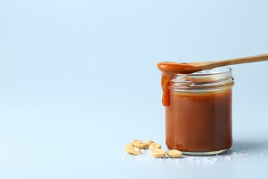 Photo of Jar of tasty salted caramel, spoon and peanuts on light blue background, space for text