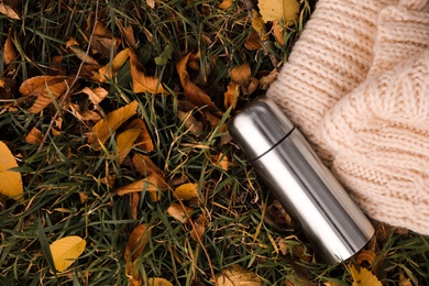 Photo of Metallic thermos and knitted scarf on green grass covered with fallen leaves outdoors, flat lay. Space for text