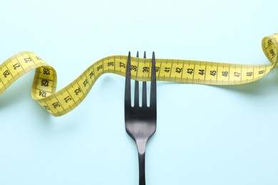 Fork with measuring tape on light blue background, top view. Diet concept