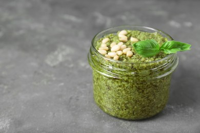 Photo of Jar with delicious pesto sauce, pine nuts and basil leaves on grey table, closeup. Space for text