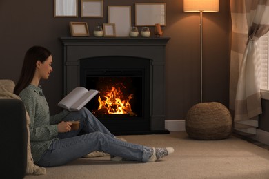 Photo of Beautiful young woman with cup of coffee reading book on floor near fireplace at home. Space for text