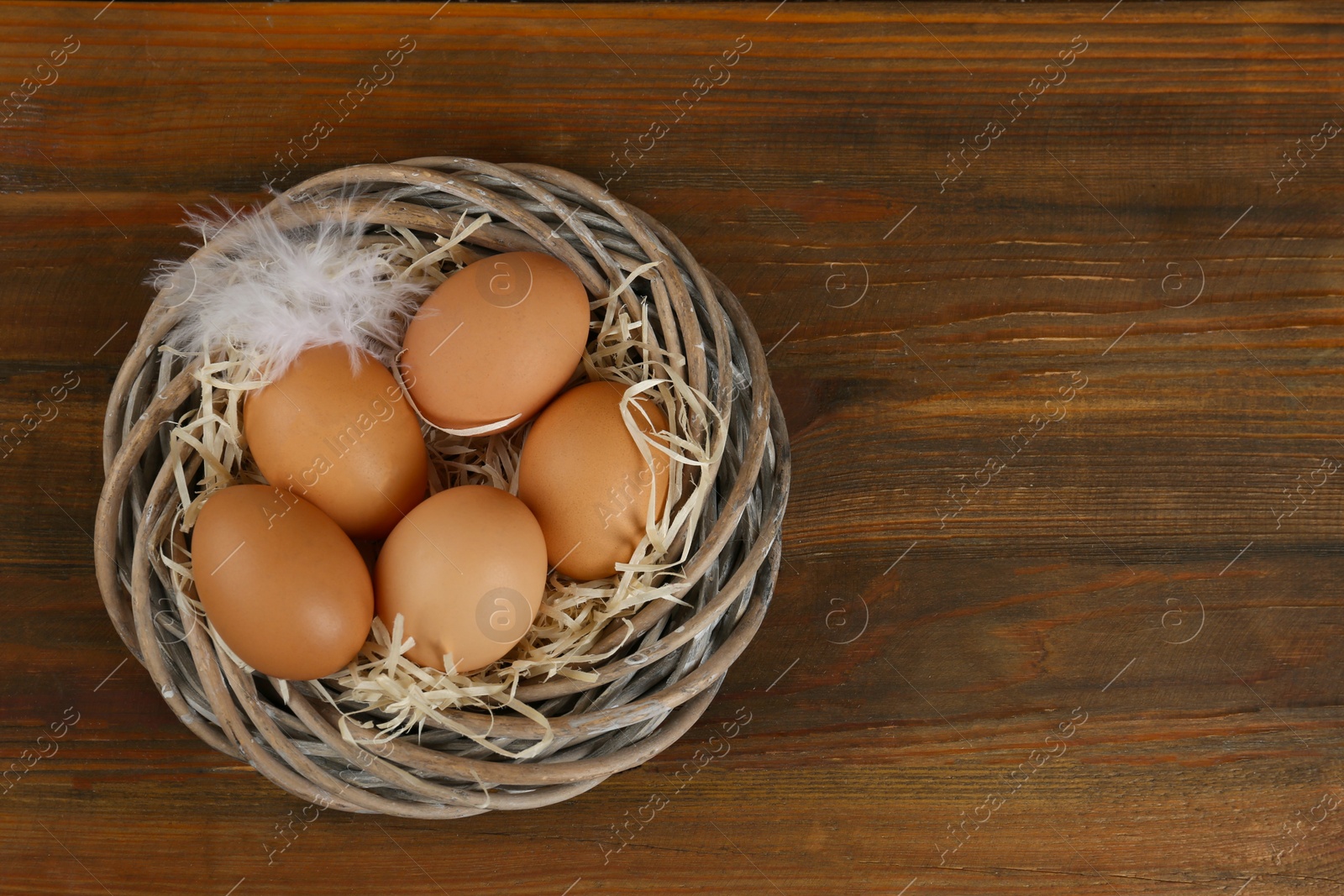 Photo of Raw chicken eggs and feather in wicker nest on wooden table, top view. Space for text