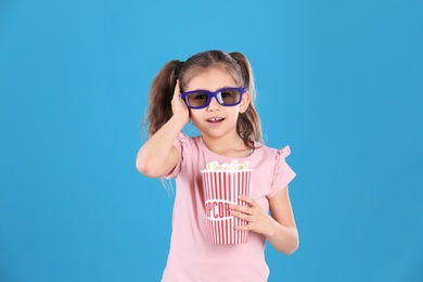 Cute little girl with popcorn and glasses on color background