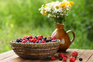 Photo of Wicker bowl with different fresh ripe berries and beautiful flowers on wooden table outdoors