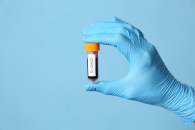 Photo of Scientist in protective gloves holding test tube with blood sample and label Covid-19 on light blue background, closeup. Space for text