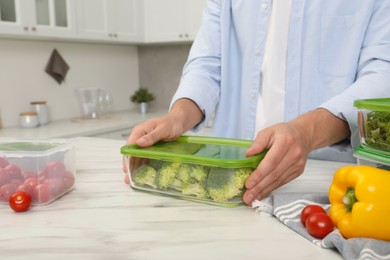Photo of Man holding glass container with fresh broccoli at white marble table in kitchen, closeup. Food storage